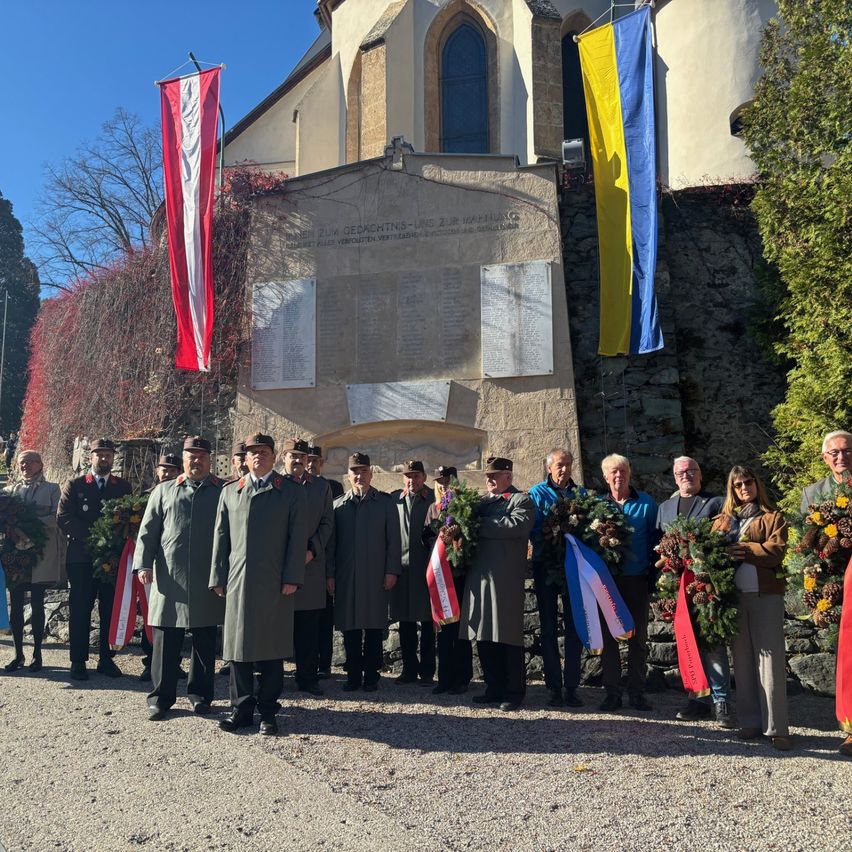 A group of people stand in front of a memorial with flags, holding wreaths and wearing uniforms.