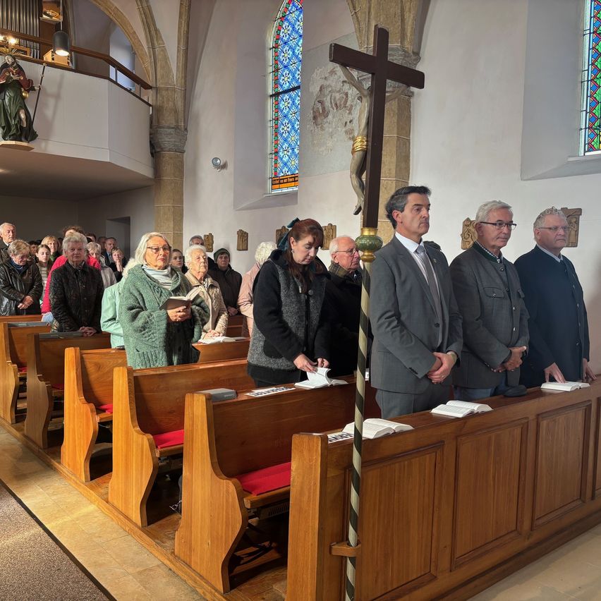 In a church, several people stand in pews, some with glasses and holding books. A large cross stands in the foreground.