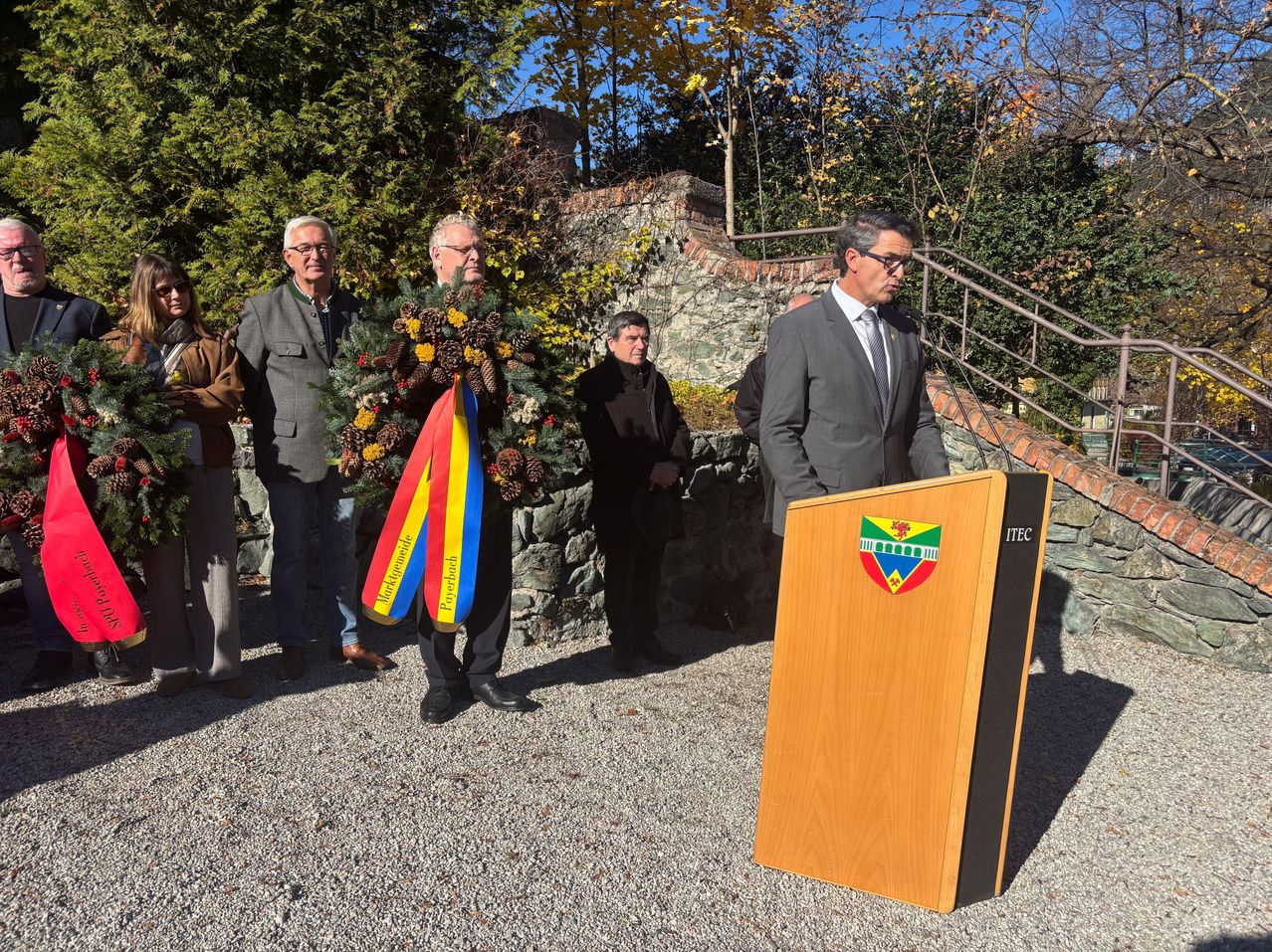 A man is giving a speech at a podium while others stand behind him. A man is holding a wreath and a flag.