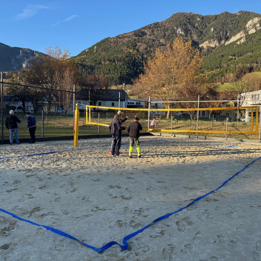People play volleyball on a sandy court with mountains in the background. The net is yellow, and the court is surrounded by a fence.