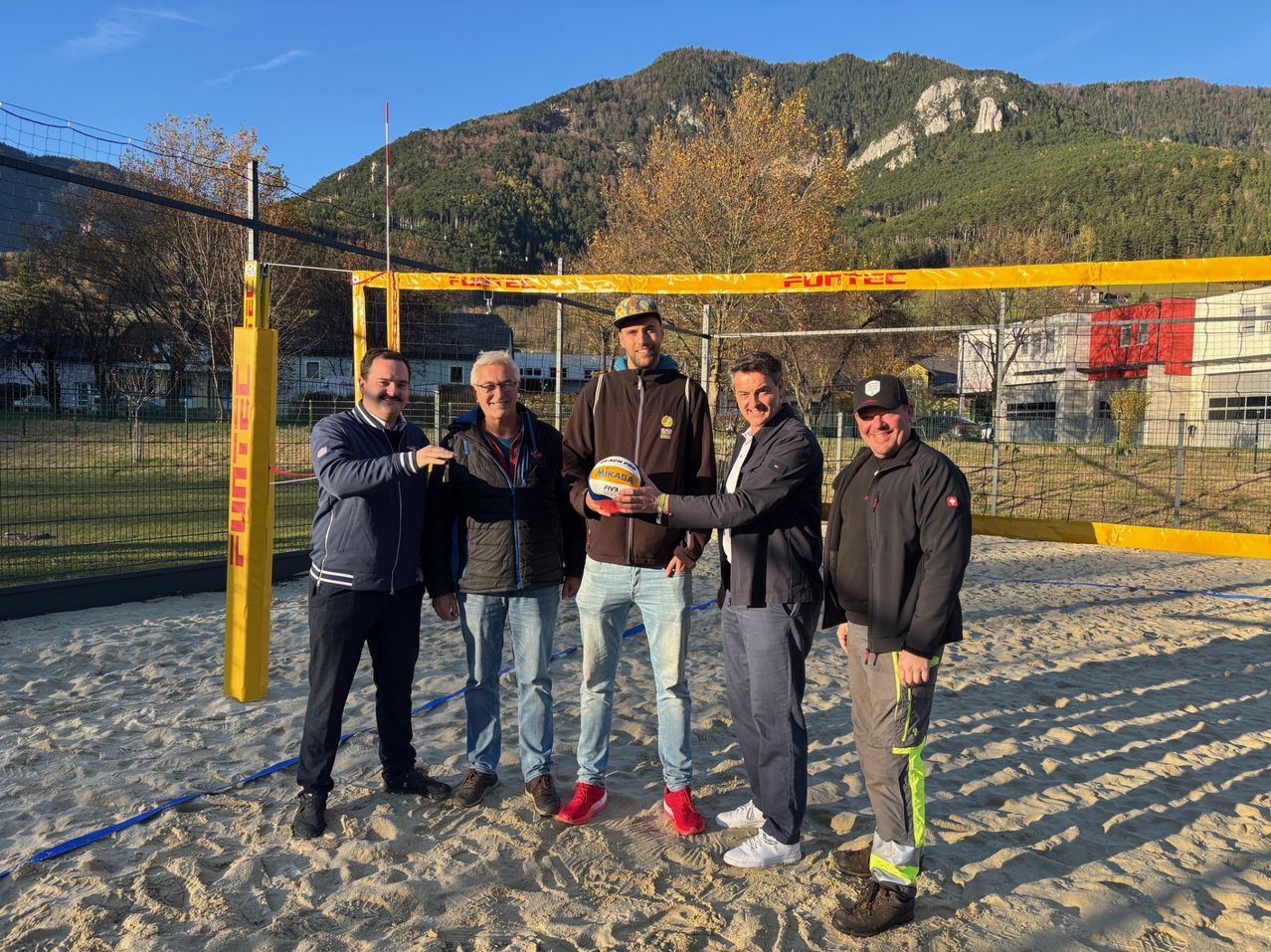 Five men stand on a beach, smiling for a photo. A volleyball net is behind them, and a mountainous backdrop. They are dressed in winter clothing.