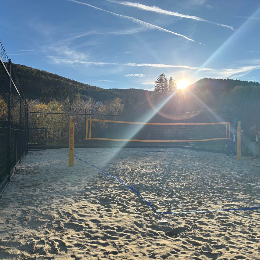 A beach volleyball court set up in a sandy area, surrounded by a metal fence. Two people are standing in the shadows. The sun is setting in the background with mountains.