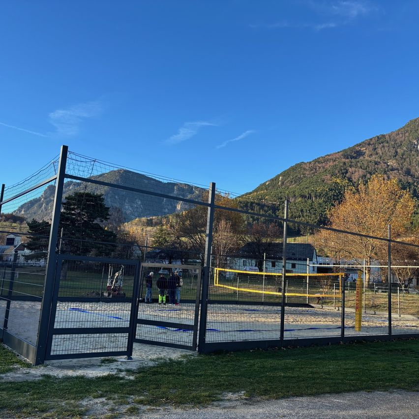 A fenced outdoor volleyball court with a net, surrounded by trees and mountains. A few people are standing near the net.