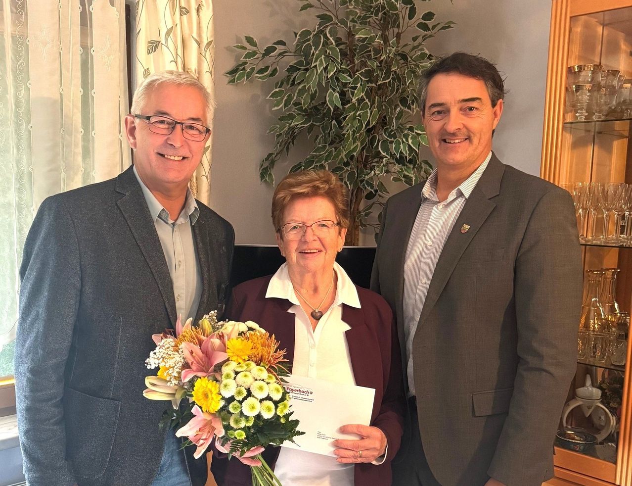 Three people stand together indoors, smiling. A woman holds a bouquet of flowers and a paper. The man on the left wears glasses and a suit. The other man on the right wears a suit with a pin. Behind them is a large plant.