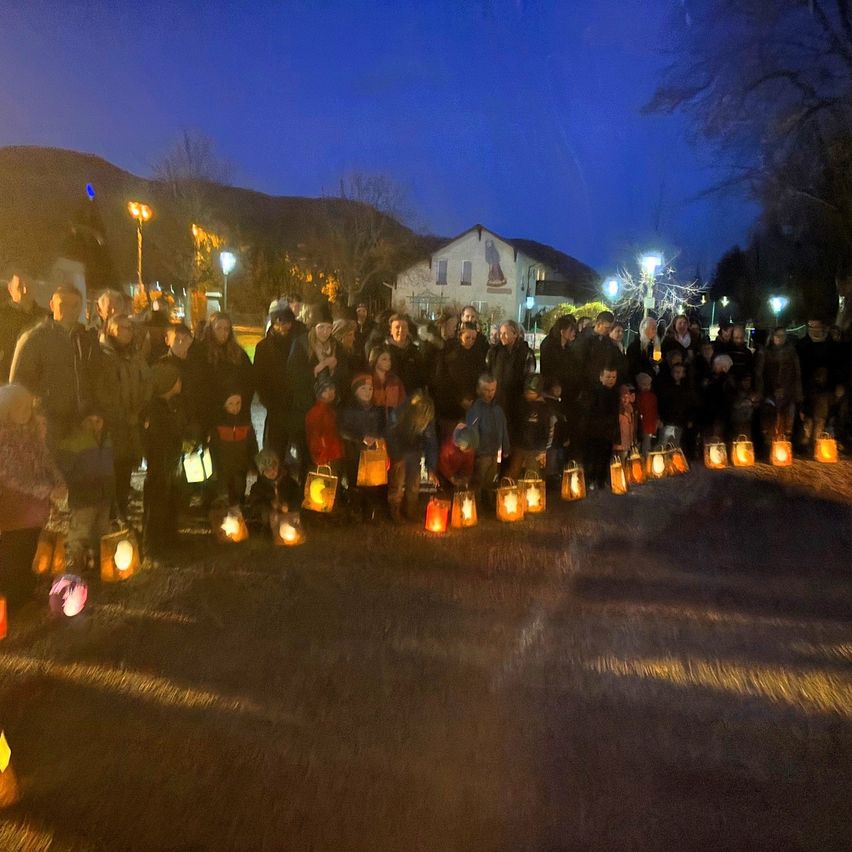 A large group of people gather at night, holding lit lanterns, with mountains and buildings in the background.