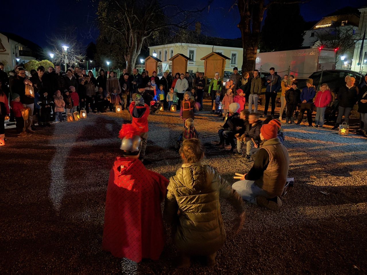 A nighttime event in a park with people dressed in costumes, holding lanterns. Some are sitting in a circle, while others stand behind them. Trees and streetlights are in the background.