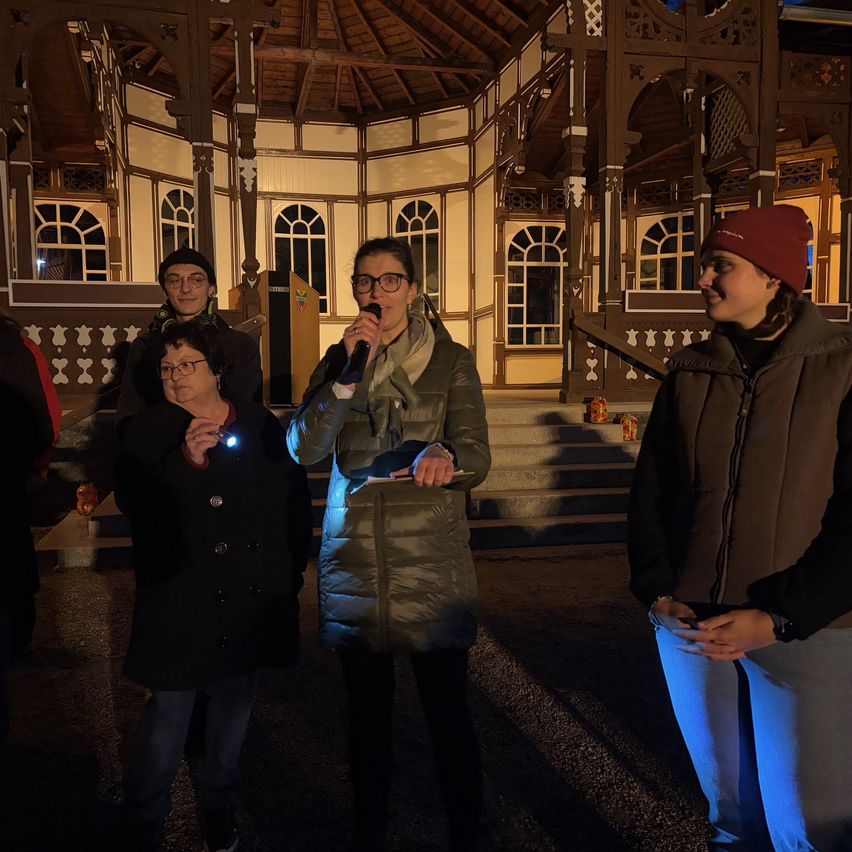 At night, a woman with a microphone stands in front of a building with arched windows and a wooden structure. Three people stand behind her.