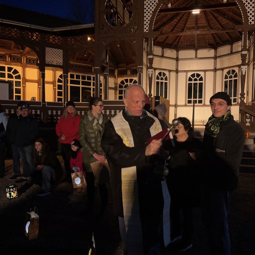 A priest holds a bell outside a gazebo at night. Several people stand and sit around him. One woman holds a cup, and a man wears a beanie.