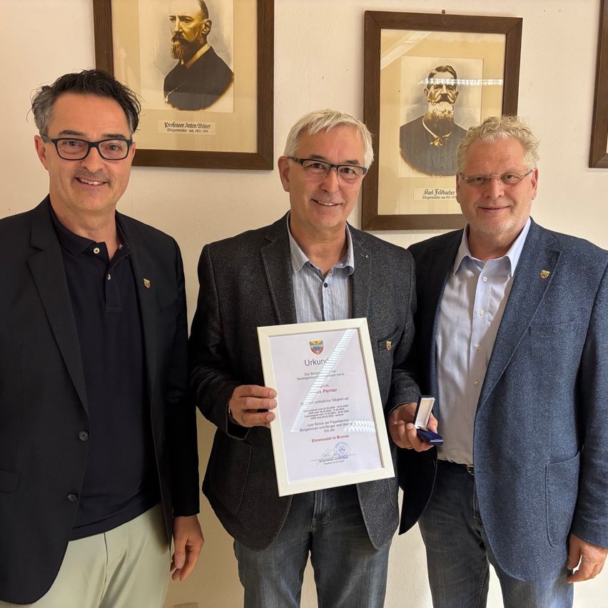 Three men in suits stand smiling in front of a wall with framed portraits. The middle man holds a certificate.