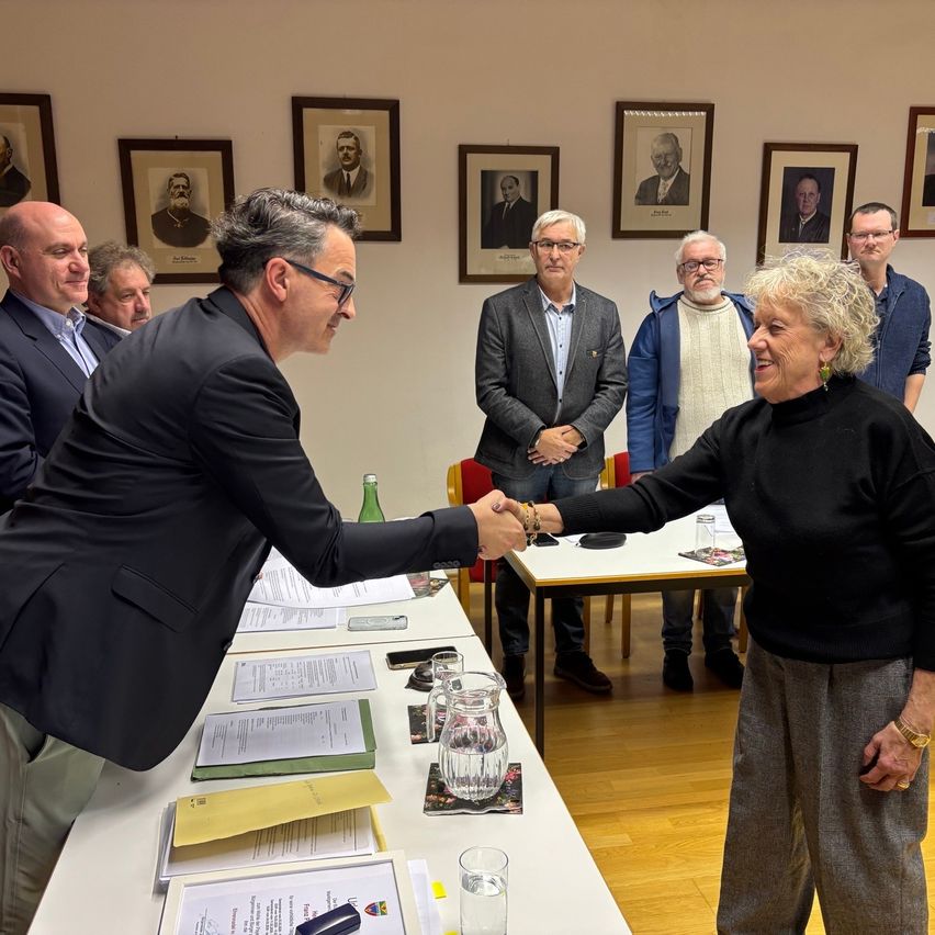 A man and a woman shake hands at a table in a meeting room. Several people stand behind them, watching. Papers, glasses, and a book are on the table. Picture frames hang on the wall.