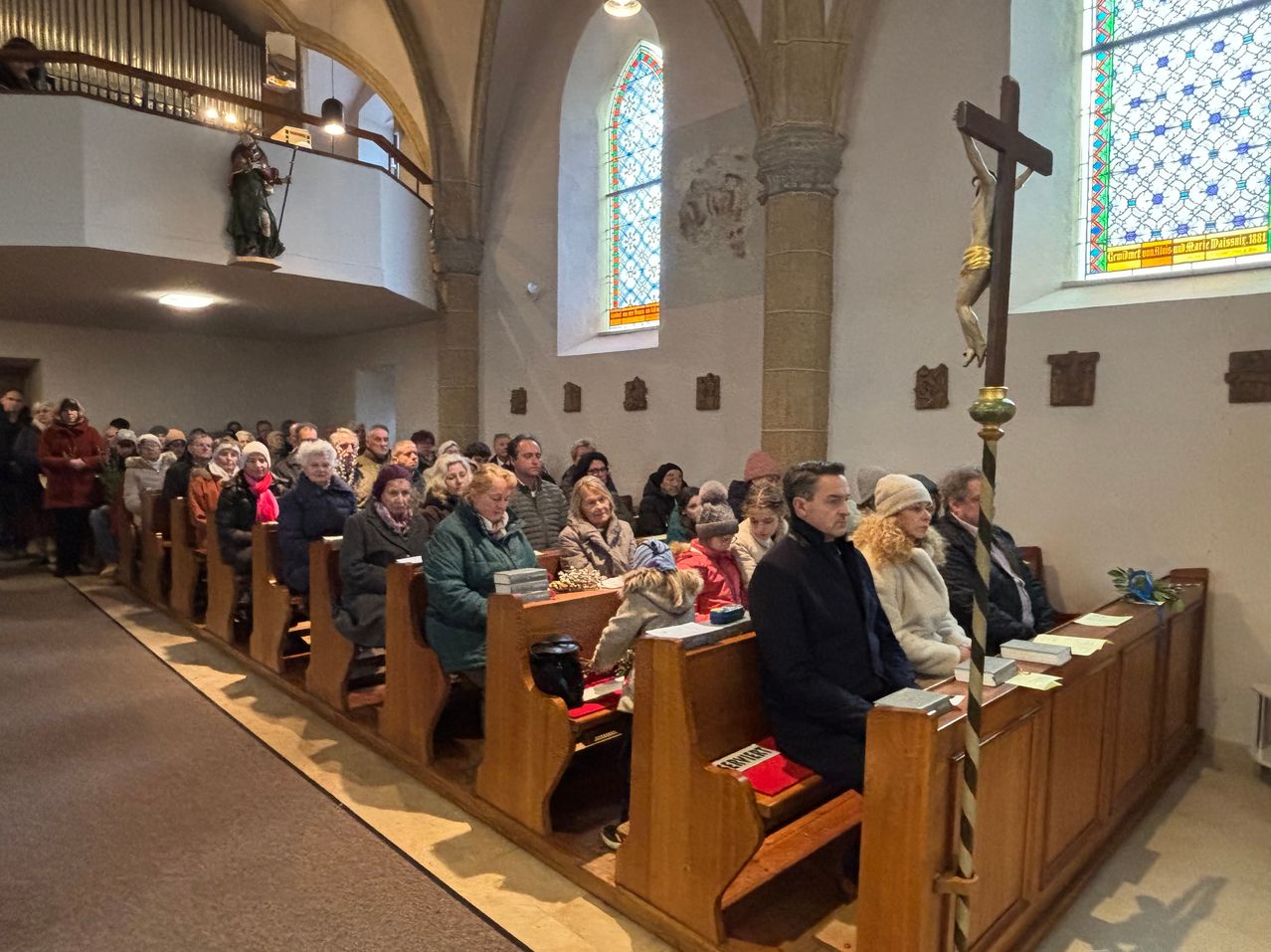 A congregation sits in pews inside a church. A crucifix stands behind the pews, and colorful stained glass windows are on the walls.