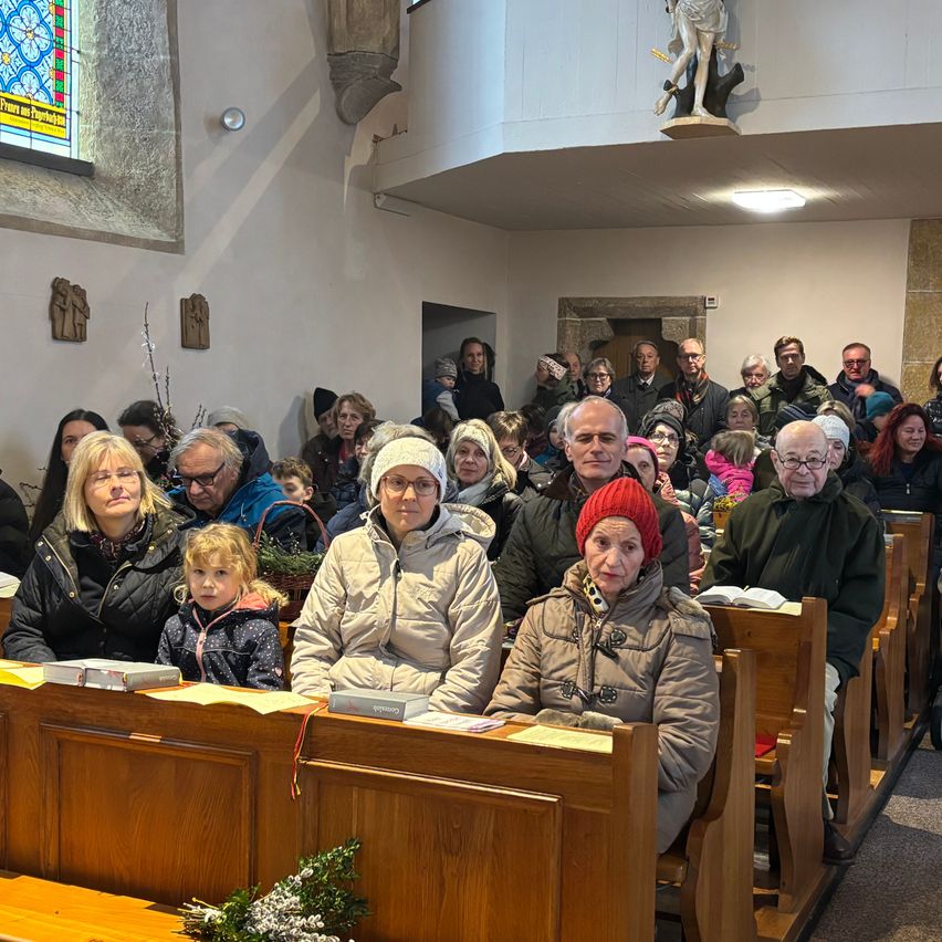 A large congregation gathers in a church, sitting on wooden pews, with a stained glass window and statue in the background.