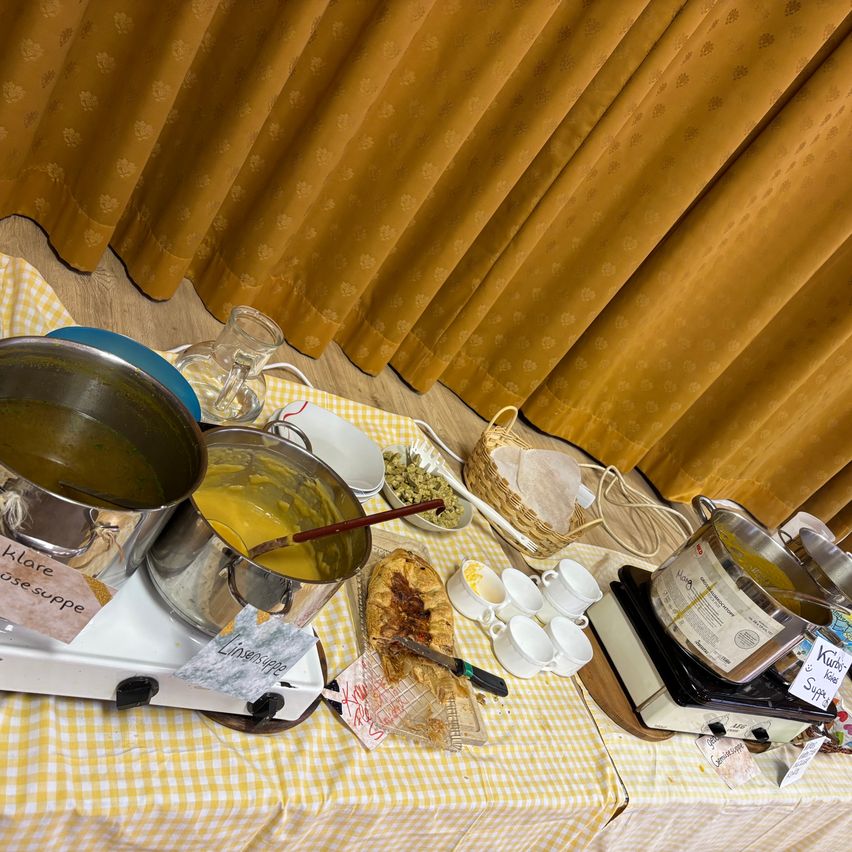 A table with several pots and pans holding different foods. It is a yellow table with a checkered tablecloth. There is a curtain in the back.