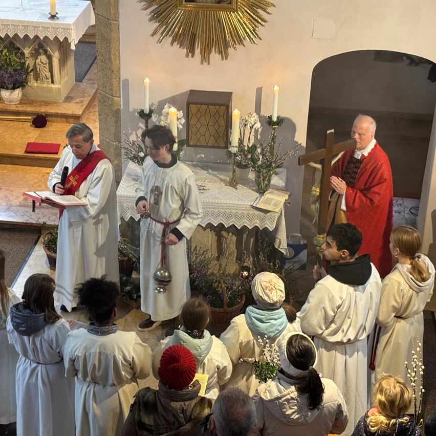A religious ceremony is taking place in a church. Two priests are standing at the altar with candles. A man is holding a book and a microphone. The congregation is dressed in white robes.