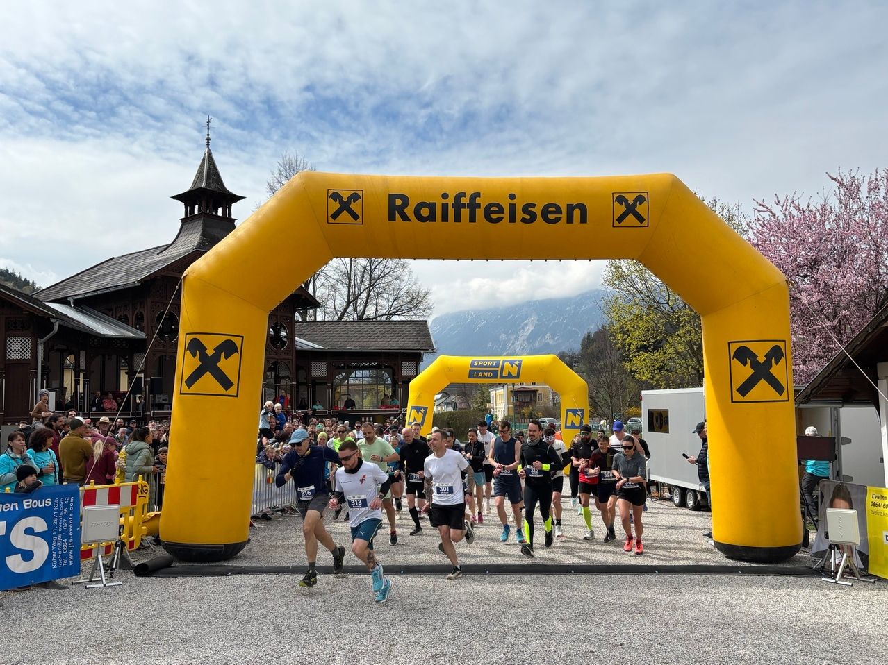 Runners pass under a yellow inflatable arch labeled 'Raiffeisen', surrounded by spectators, buildings, and mountains.