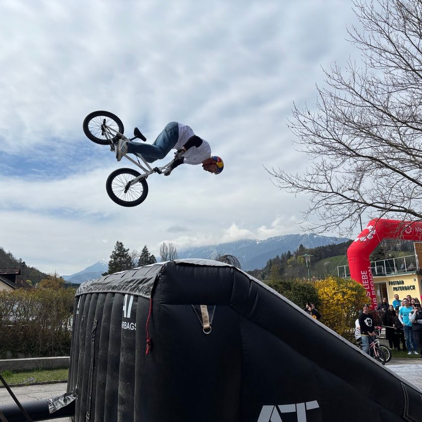 A cyclist performs a stunt by flipping a bicycle in the air while wearing a helmet. The event is held outdoors with spectators watching from the side. The background shows mountains and a cloudy sky.