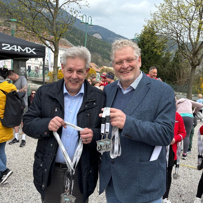 Two men holding medals stand smiling at an outdoor event. They wear winter coats and are surrounded by people and trees. A canopy with the text 24MX is behind them.