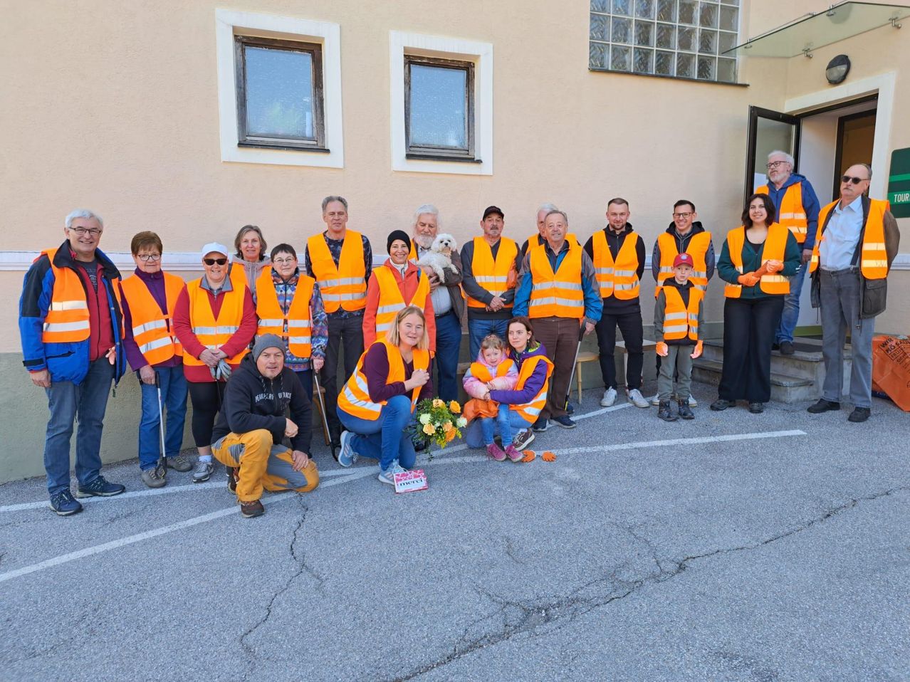 A group of people in orange vests and hats stand in front of a building. A woman holds a flower bouquet, and a child holds a small orange. Some people wear glasses and hats.