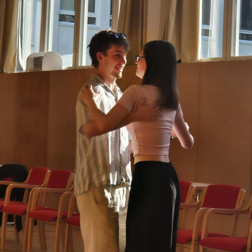 A man and a woman are dancing in a room with red chairs and windows with curtains.