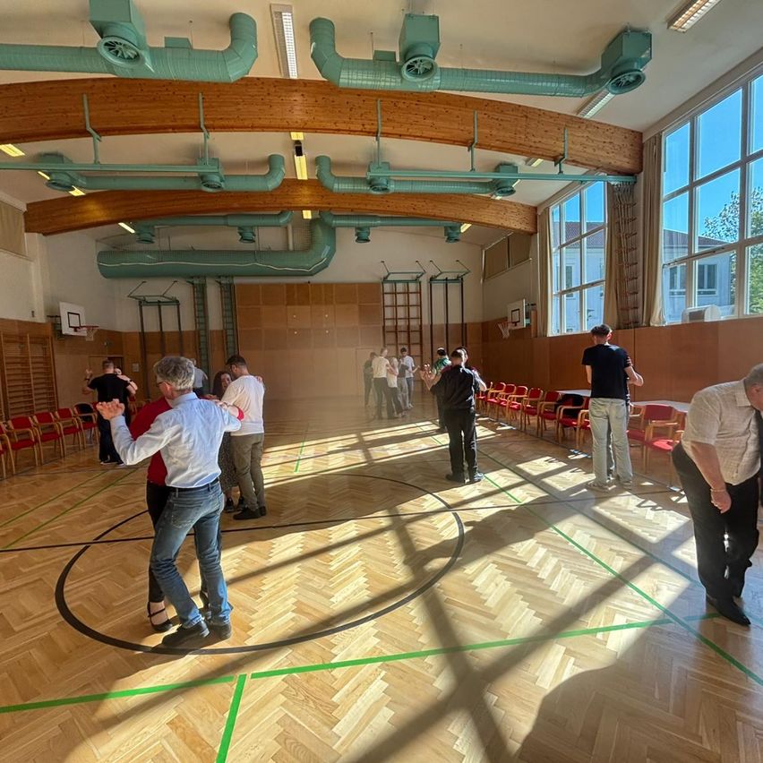A group of people dance in a gymnasium. The room is spacious with wooden floors and large windows. Some individuals are standing near chairs and a table.