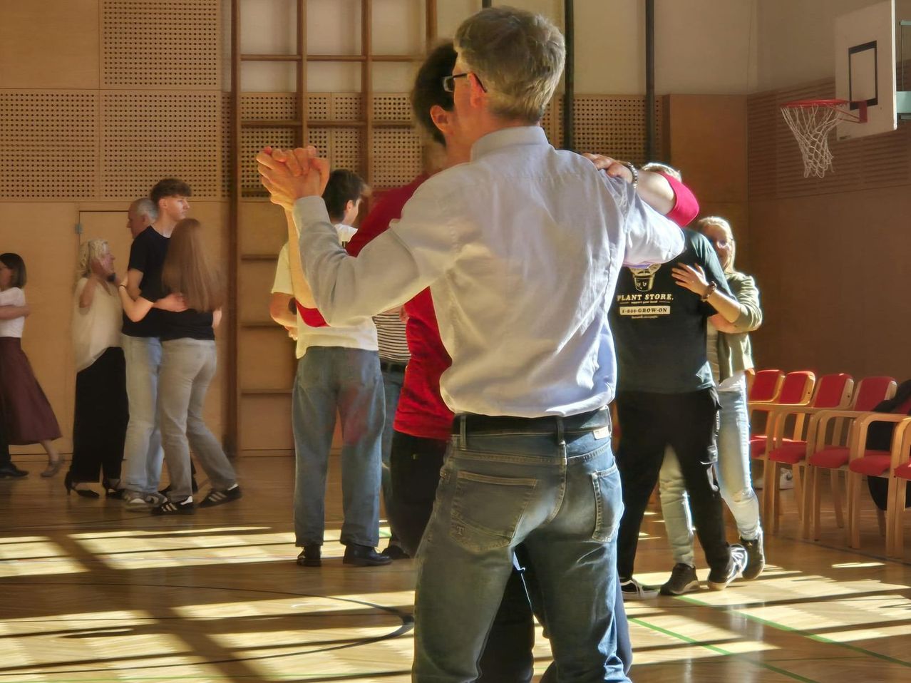 A group of people dancing in a room with a wooden wall, a basketball hoop, and a red chair.