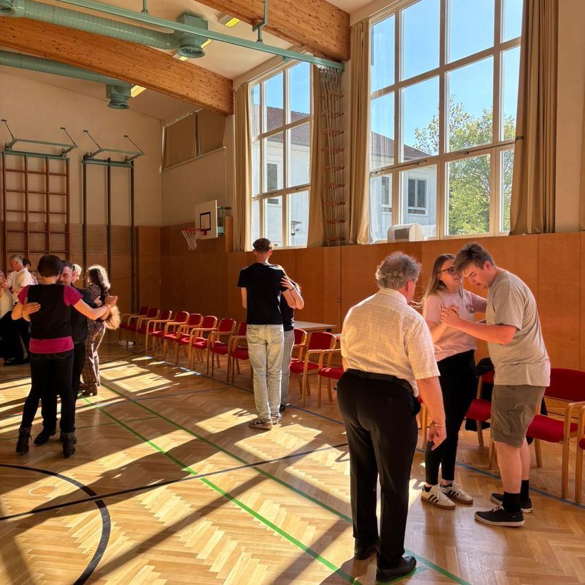 People practice dancing in a gymnasium with wooden floors and large windows. Some are standing, and a few are in pairs.