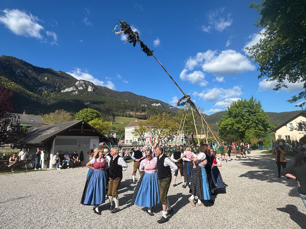 A group of people dressed in traditional costumes dance around a tall pole on a gravel surface with mountains in the background.