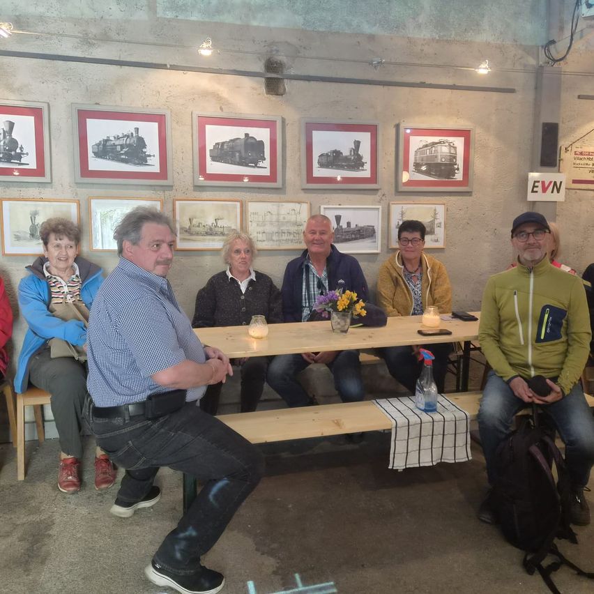 A group of people sit around a wooden table with flowers and candles in a room with framed black-and-white train pictures on the wall.