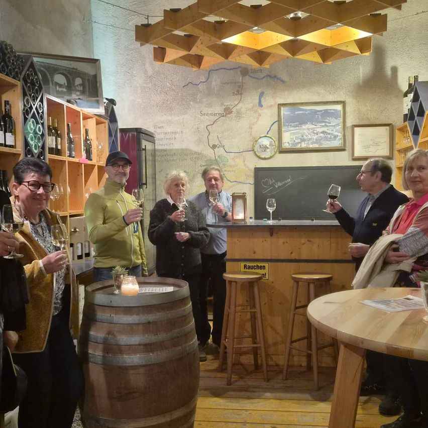 A group of people are in a wine tasting room, holding wine glasses. They are standing around a barrel and a table with wine bottles on the shelves.
