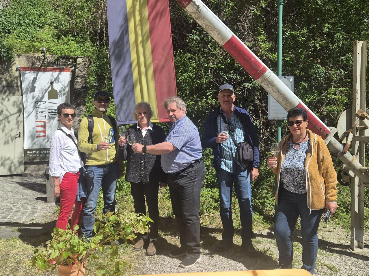 A group of people, including men and women, are standing in front of a flag and holding wine glasses. They seem to be posing for a photograph.