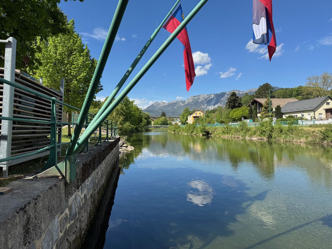 A serene river with flags and mountains in the distance. Trees and houses line the riverbanks.