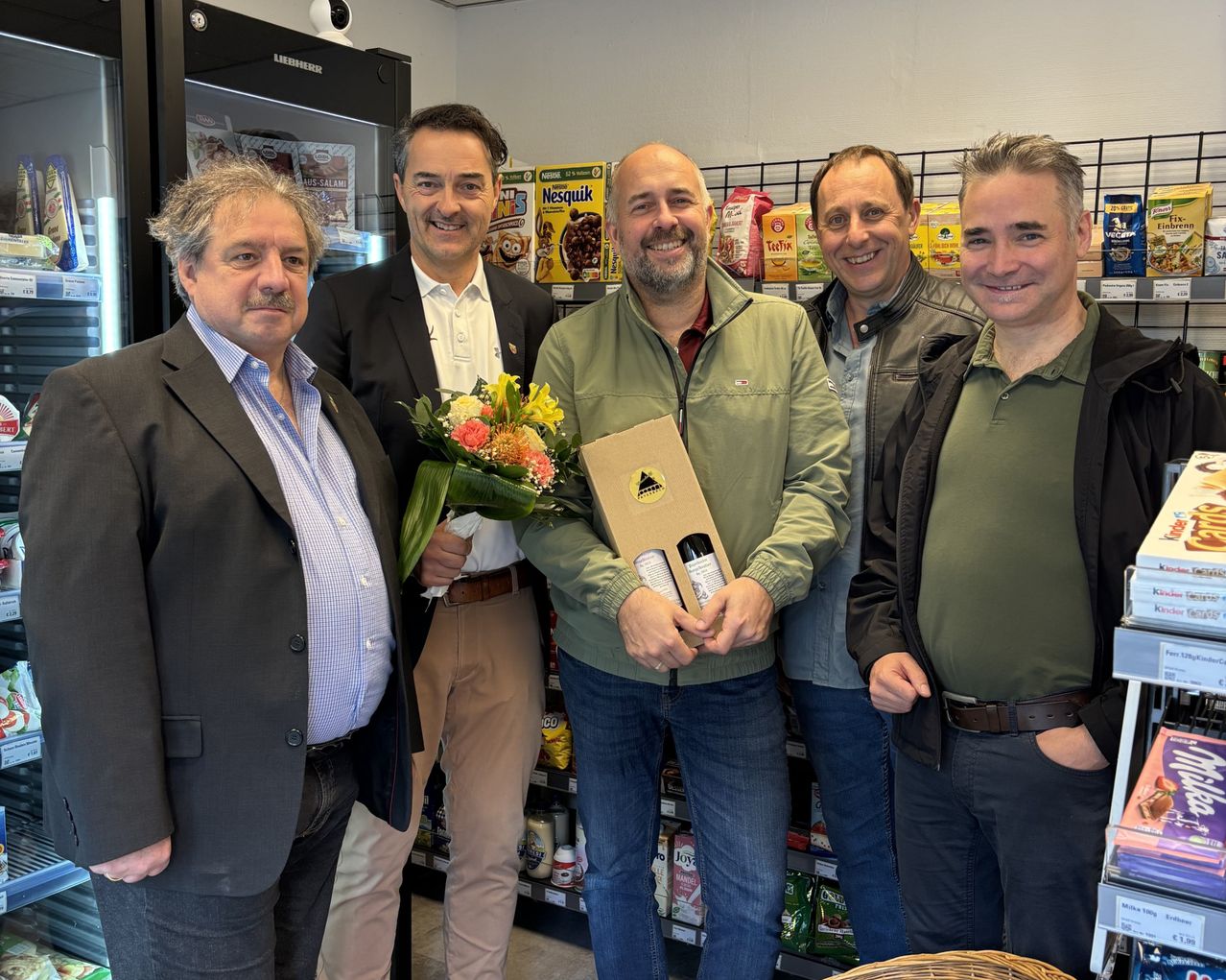 Fünf Männer stehen lächelnd vor einem Geschäft und posieren für ein Foto. Ein Mann hält einen Blumenstrauß und eine Geschenkbox in der Hand. Hinter ihnen ist ein Regal mit verschiedenen Produkten zu sehen, darunter Müsli und Snacks.