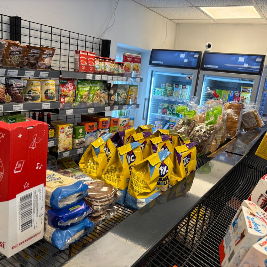 Ein Lebensmittelgeschäft mit verschiedenen Produkten zur Schau, darunter verpackte Snacks, Brot und Tiefkühlprodukte in Kühlschränken. Die Vorderseite hat eine Kasse mit verschiedenen Snacks und verpackten Waren.