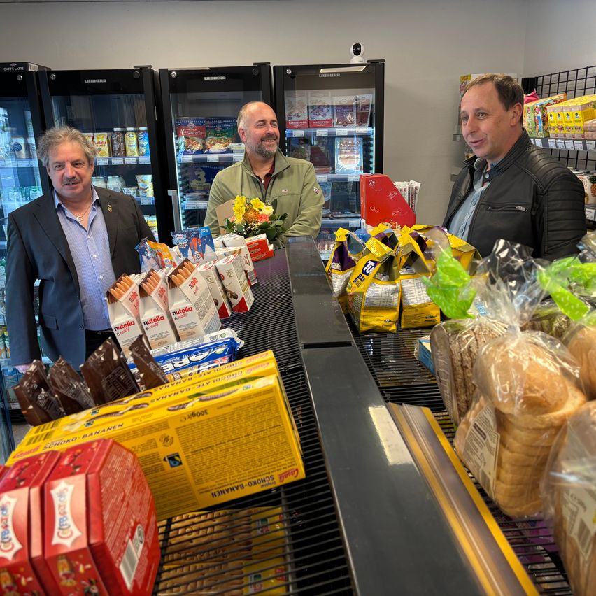Drei Männer stehen in einem Geschäft mit Regalen im Hintergrund. Es sind verschiedene Lebensmittel ausgestellt, darunter Brot, Kekse und ein Blumenstrauß.