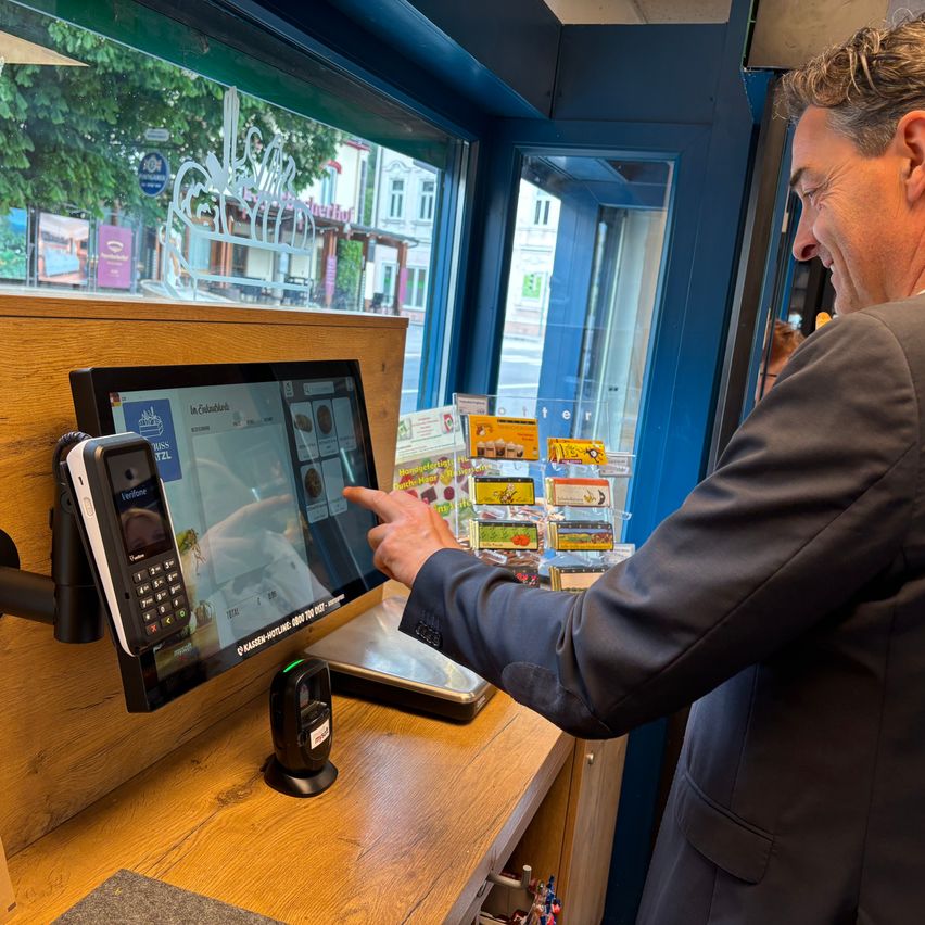 Ein Mann in einem Anzug benutzt ein Touchscreen-Gerät, das an einer hölzernen Theke in einem Geschäft montiert ist. Hinter ihm zeigt eine Glastür verschiedene Artikel an.