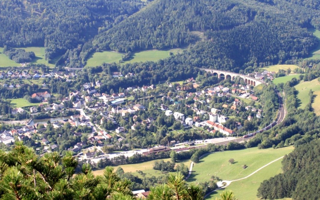 Luftaufnahme einer kleinen Stadt umgeben von üppigem Grün und Bergen. Eine Brücke überquert einen Fluss und ein Zug durchquert die Landschaft.