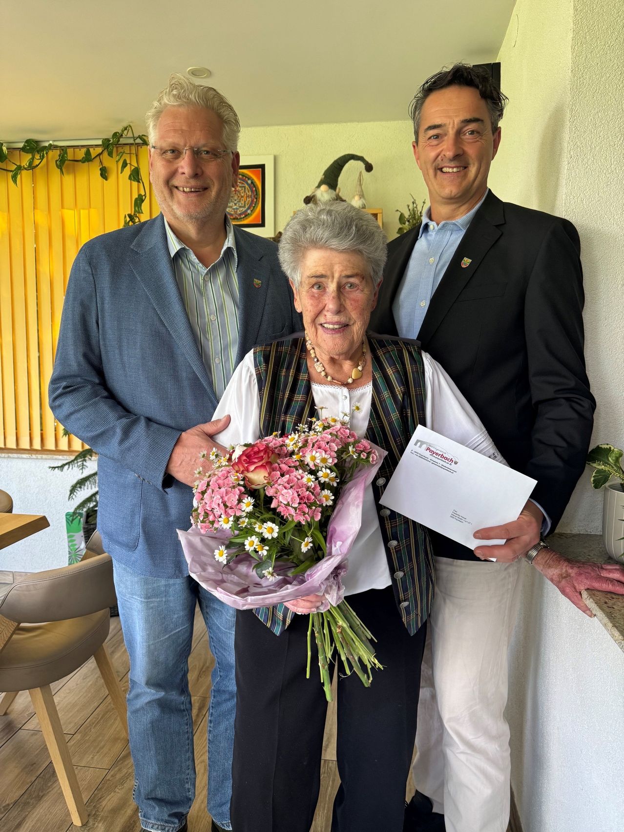Drei Männer stehen hinter einer älteren Frau, die Blumen und einen Brief in der Hand hält. Sie alle lächeln und scheinen für ein Foto zu posieren.