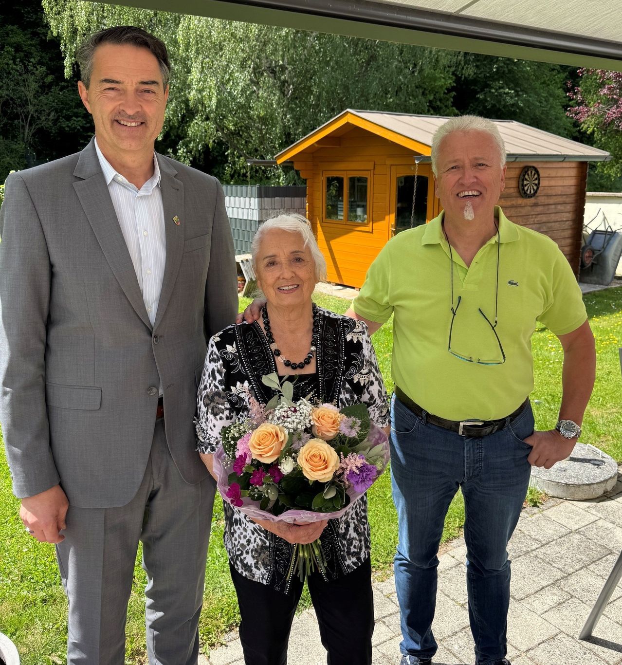 Drei Personen stehen in einem Garten. Eine Frau hält Blumen, lächelt und steht zwischen zwei Männern. Einer trägt einen Anzug, der andere ein grünes Poloshirt. Sie stehen neben einem kleinen orangefarbenen Haus.