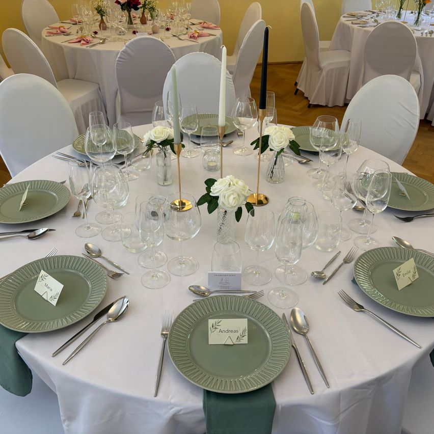 Ein formeller Esstisch, der mit weißen Tischdecken, grünen Tellern und Kerzen gedeckt ist. Besteck und Gläser sind um den Tisch herum angeordnet, mit Blumenarrangements und Namenskarten.