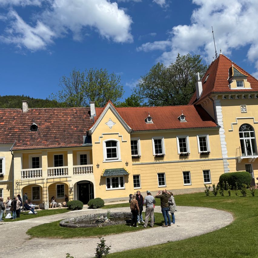 Eine Gruppe von Menschen versammelt sich vor einem gelben Schloss. Das Schloss hat ein rotes Dach und viele Fenster. Der Himmel ist blau mit Wolken.