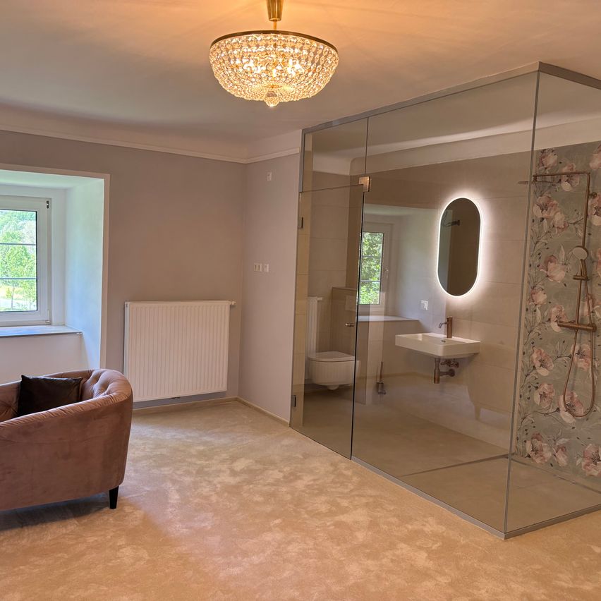 Ein modernes Badezimmer mit einer Glasdusche, einer Kristallleuchte und einem weißen Heizkörper. In der Ecke steht eine Couch.