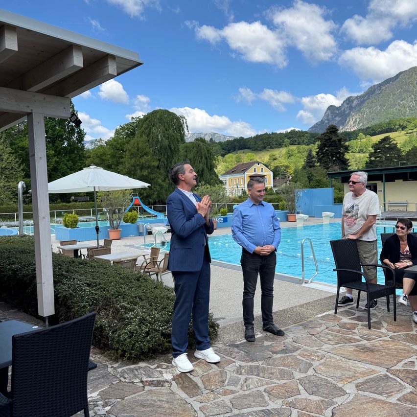 Eine Gruppe von Männern und Frauen steht am Rand eines Schwimmbeckens, zwei der Männer tragen Anzüge. Sie scheinen für ein Foto zu posieren.