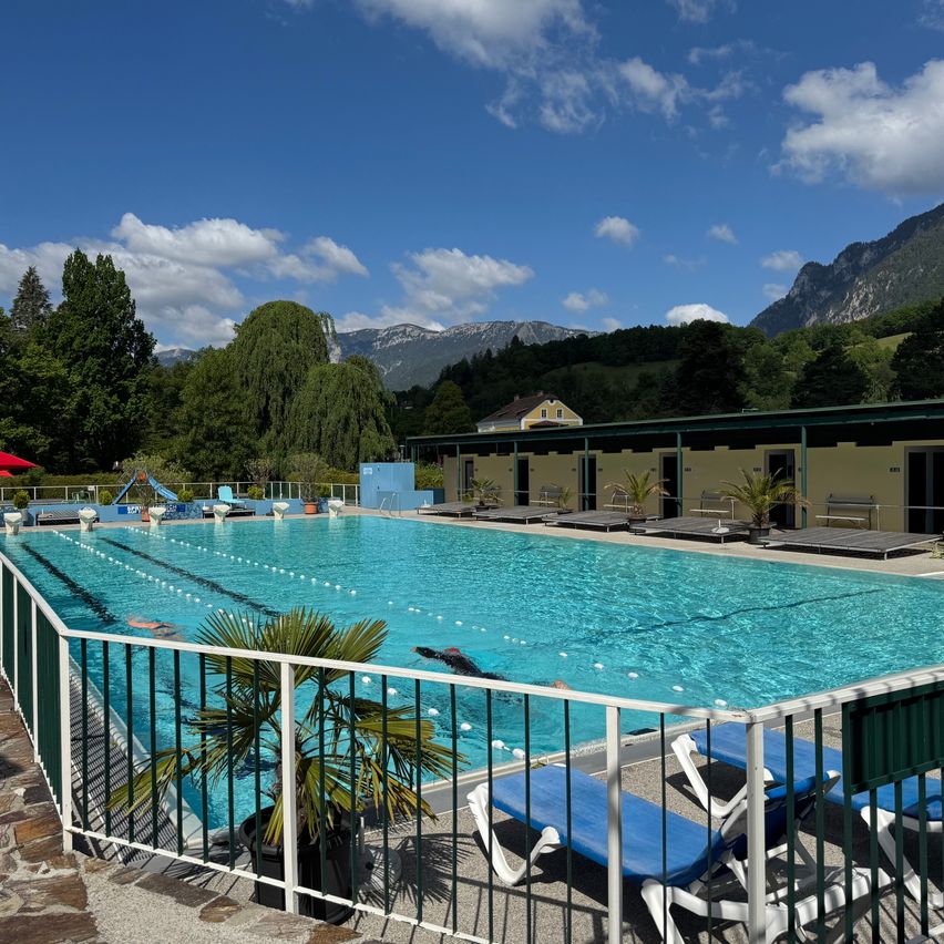 Ein Freibad mit klarem blauem Wasser, einem roten Sonnenschirm, Liegestühlen und einem Haus im Hintergrund mit Bergen.