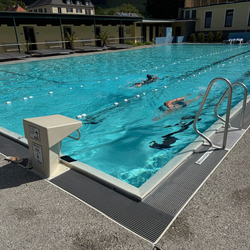 Zwei Menschen schwimmen Bahnen in einem großen Freibad. Das Becken ist von Gebäuden, Pflanzen und Liegestühlen umgeben. Ein Schwimmer trägt eine blaue Kappe.