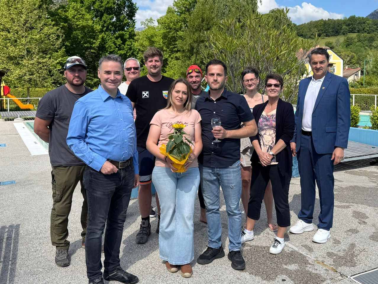 Eine Gruppe von Menschen steht lächelnd für ein Foto in einem Park. Eine Frau in der Mitte hält eine gelbe Blume. Männer und Frauen tragen Freizeitkleidung und Turnschuhe. Bäume und ein Gebäude sind im Hintergrund.