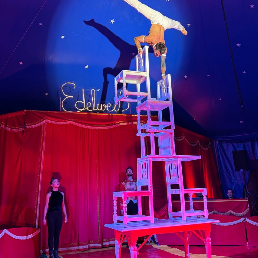 Ein Mann balanciert auf einer Stuhlpyramide im Zirkus. Er ist kopfüber und seine Hände liegen auf den Stühlen. Zwei Personen beobachten ihn von der Seite.