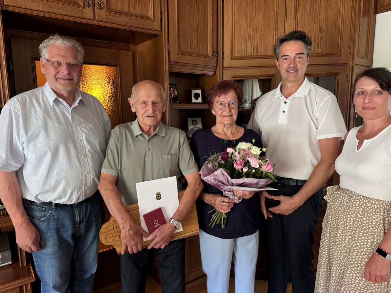 Vier Erwachsene stehen in einem Raum, zwei Männer und zwei Frauen, lächeln und posieren für ein Foto. Der Mann auf der linken Seite hält ein Buch und eine rote Mappe. Die Frau in der Mitte hält einen Blumenstrauß.
