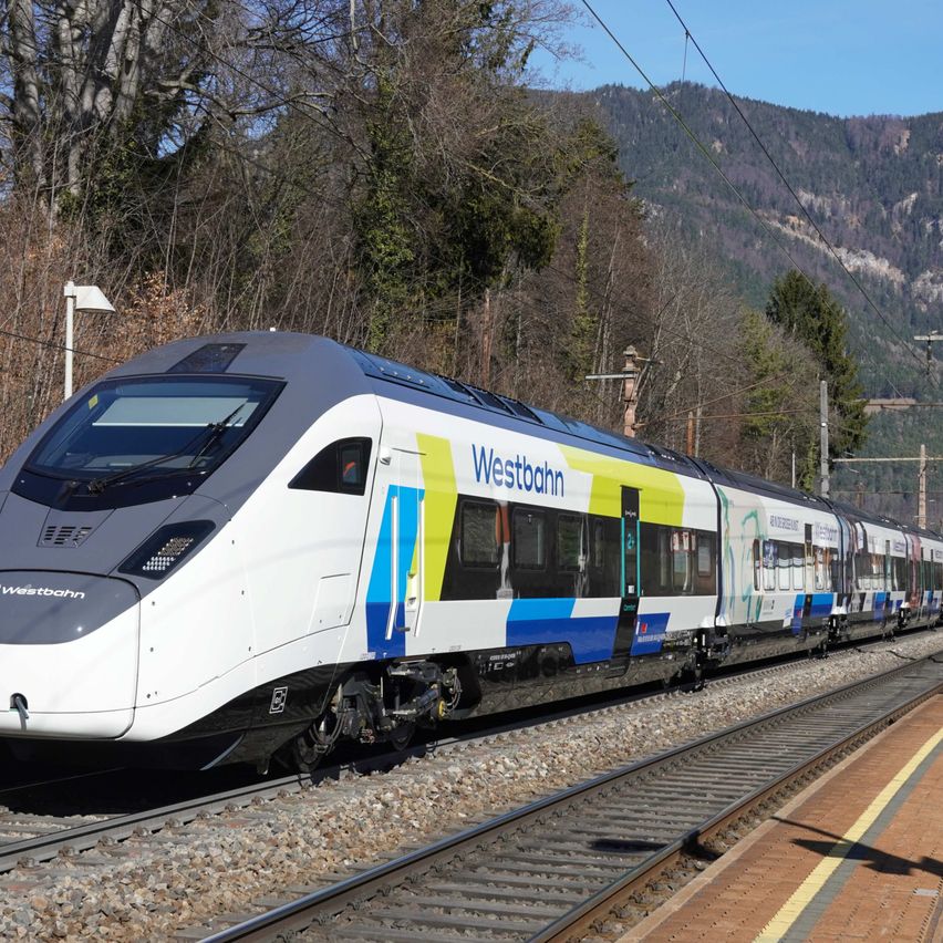 Ein Westbahn-Zug fährt auf einer Strecke in einem bergigen Gebiet mit grünen Bäumen, unter einem klaren blauen Himmel.