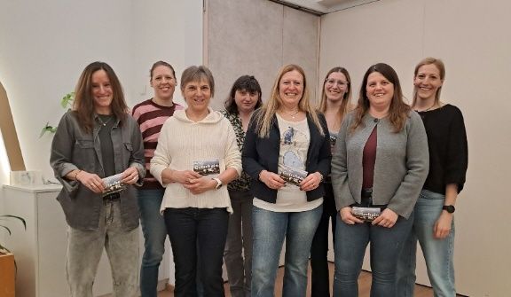 Eine Gruppe von Frauen steht lächelnd zusammen und hält Bücher in einem Raum mit weißer Wand und Holzboden.