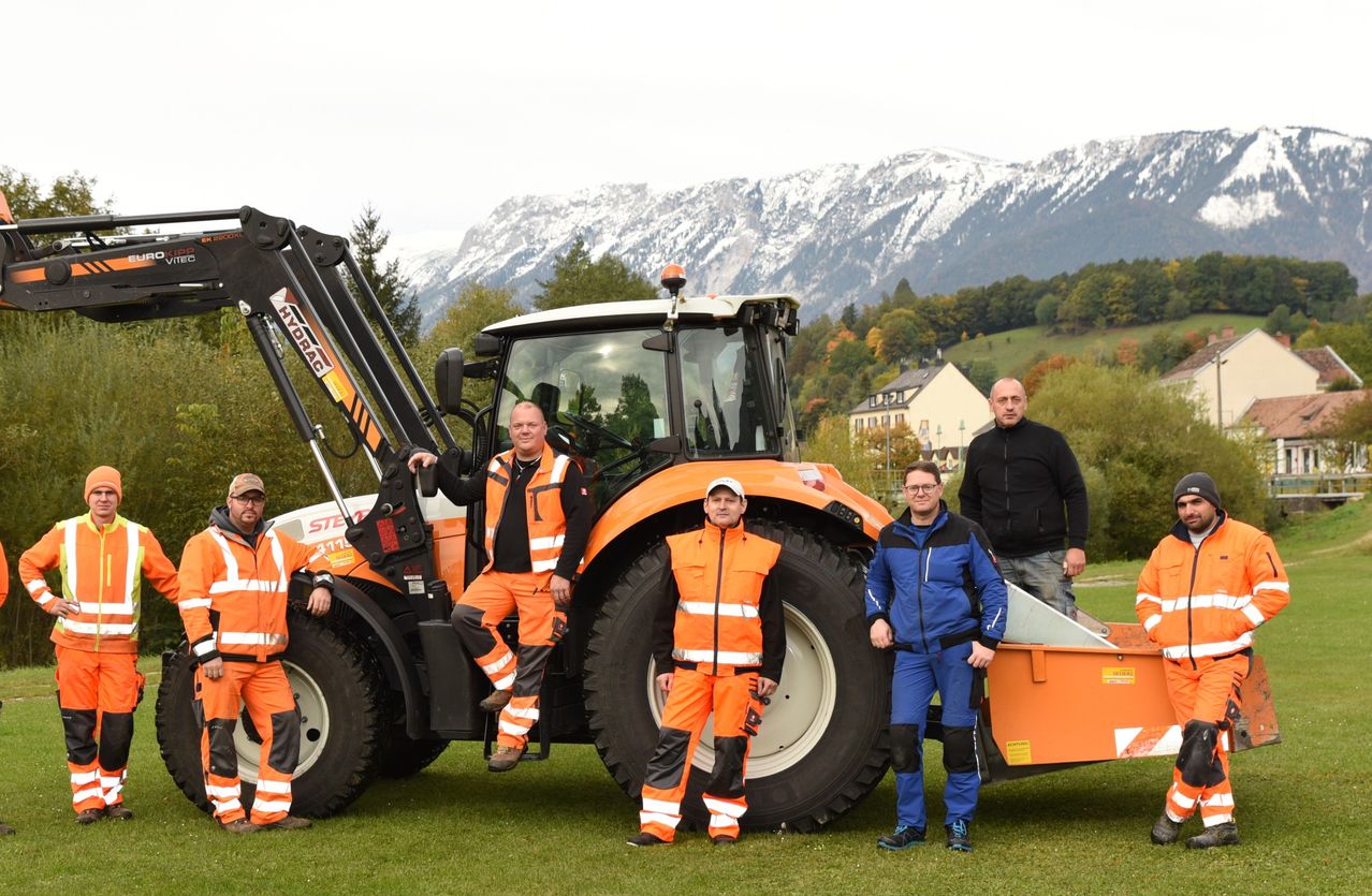 Fünf Männer in orangefarbenen Sicherheitsanzügen stehen vor einem großen orangefarbenen Traktor mit verschneiten Bergen im Hintergrund.